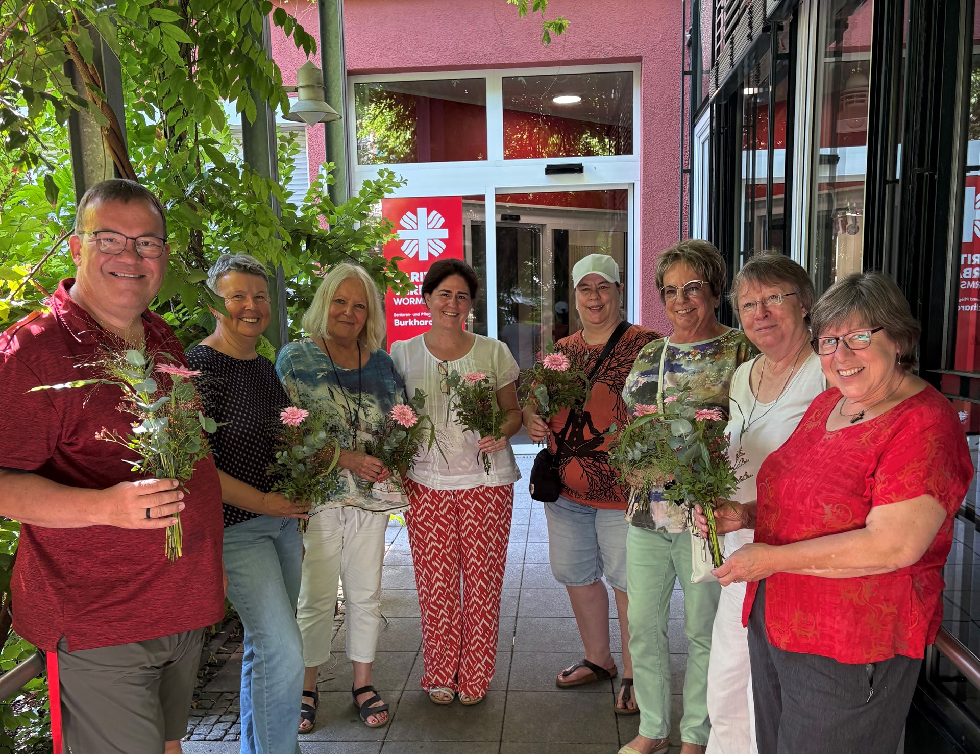 Acht Erwachsene stehen vor dem Eingang einer Caritas-Einrichtung und halten selbst gebundene Blumensträuße in den Händen. Die Gruppe lächelt in die Kamera. Im Hintergrund sind Pflanzen, der Eingangsbereich des Gebäudes und ein Caritas-Schild zu sehen. Die Aufnahme vermittelt Gemeinschaft und ehrenamtliches Engagement.