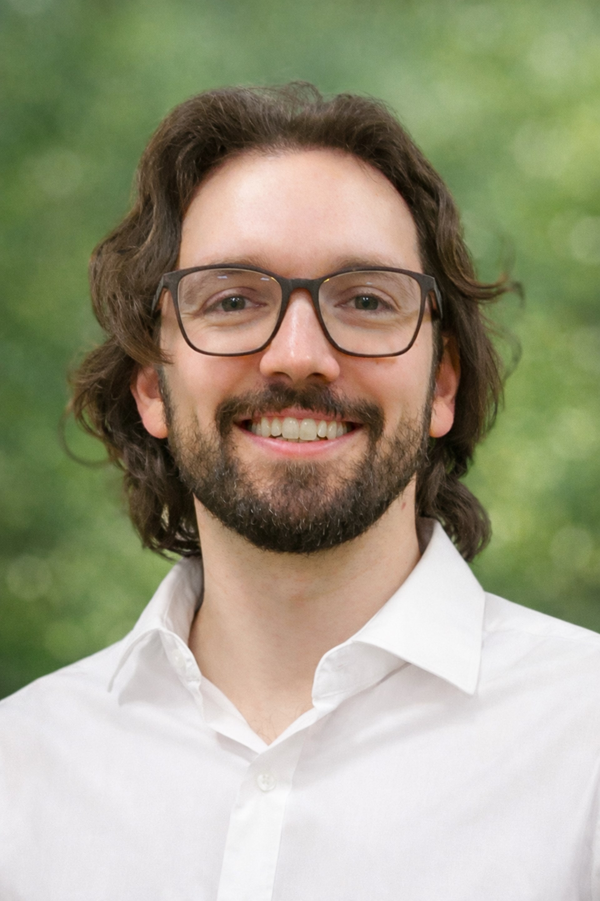 Porträt eines lächelnden Mannes mit Brille, Bart und schulterlangem Haar vor hellem Hintergrund.