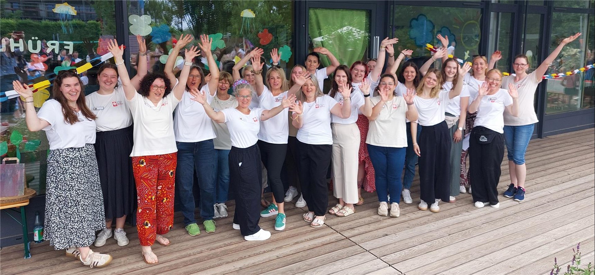Team-Foto des Familienzentrums Kita Ache Noah - viele Frauen stehen auf einer Terrasse vor einer geschmückten Fensterfront und winken gut gelaunt in die Kamera