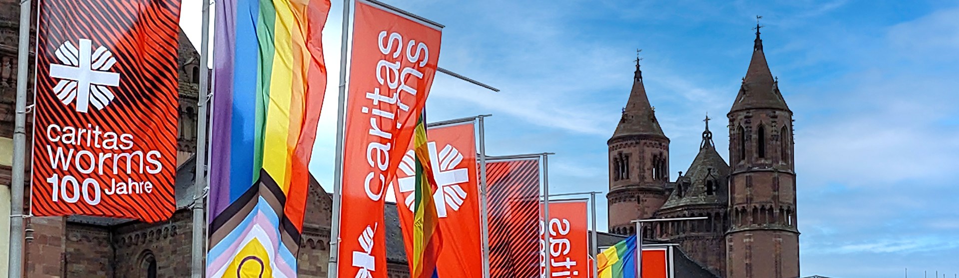 Mehrere Fahnen mit dem Schriftzug „caritas worms 100 Jahre“ und Regenbogenflaggen wehen vor dem Wormser Dom. Im Hintergrund sind die Türme der Kathedrale unter blauem Himmel zu sehen.