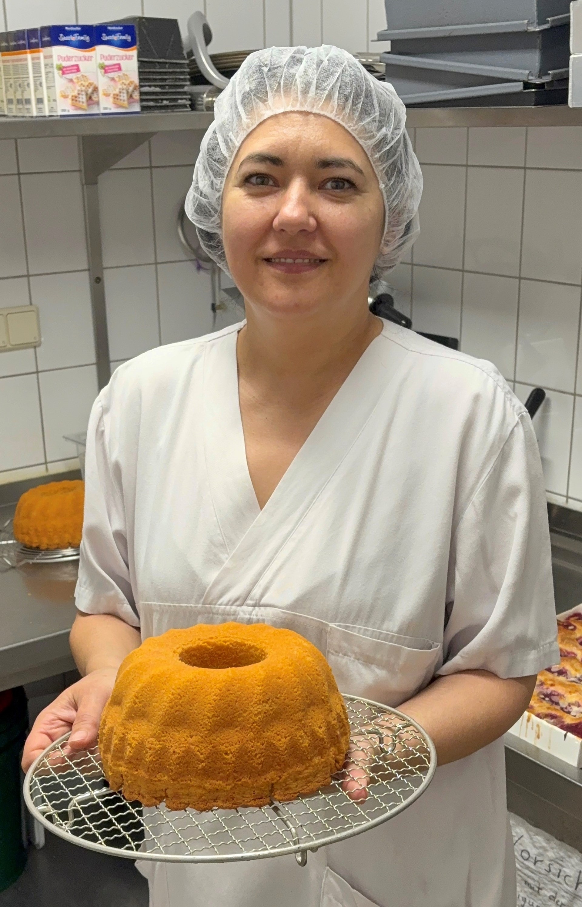 Mitarbeiterin einer Großküche in weißer Arbeitskleidung und Haarnetz hält einen frisch gebackenen Gugelhupf auf einem Kuchengitter. Sie steht in einer gewerblichen Küche mit Edelstahlflächen, Backformen und weiteren Backwaren im Hintergrund. Die Mitarbeiterin blickt lächelnd in die Kamera.