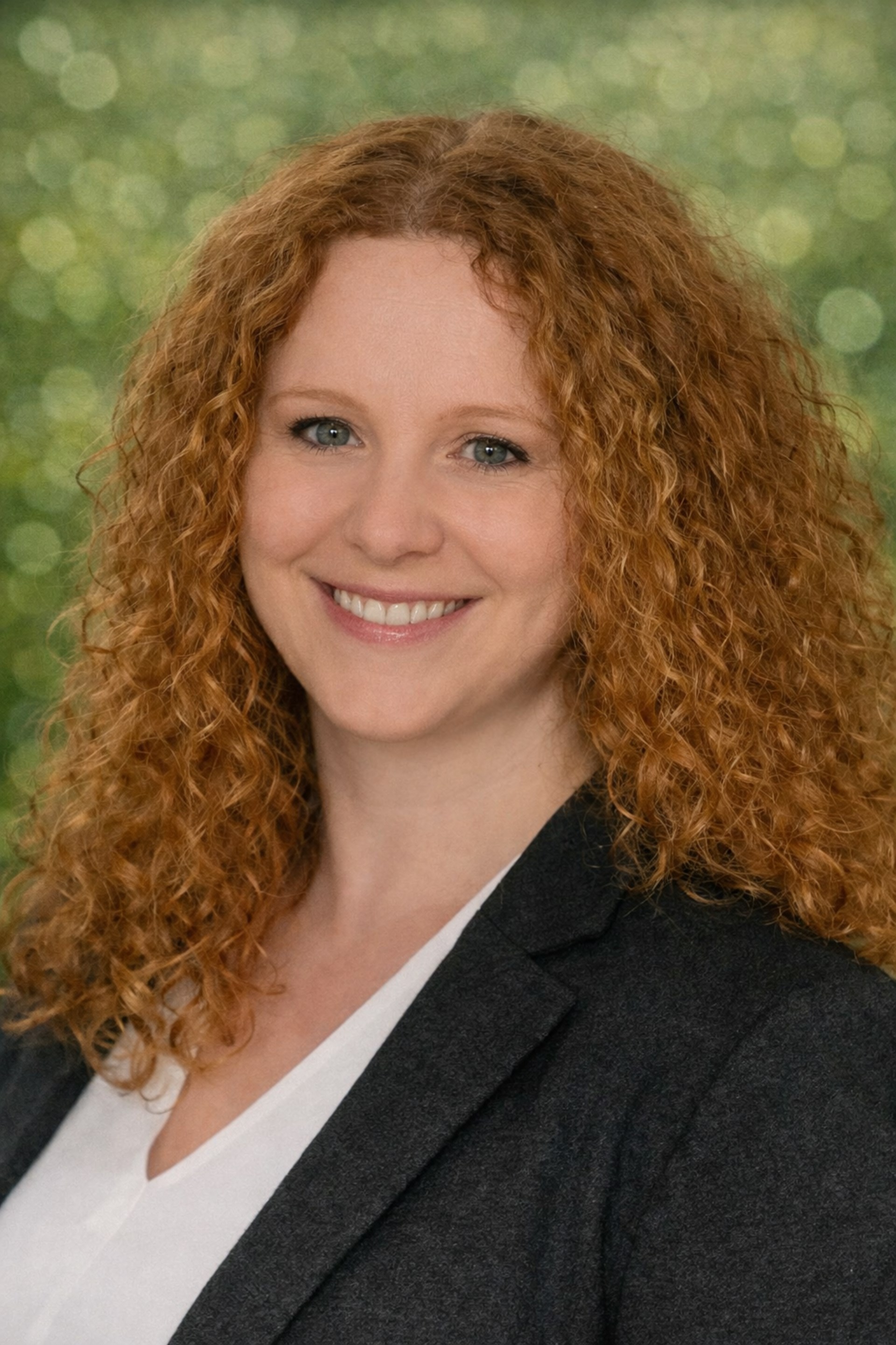 Porträt einer freundlich lächelnden Frau mit roten Locken vor einem unscharfen grünen Hintergrund. Sie trägt eine weiße Bluse und einen schwarzen Blazer