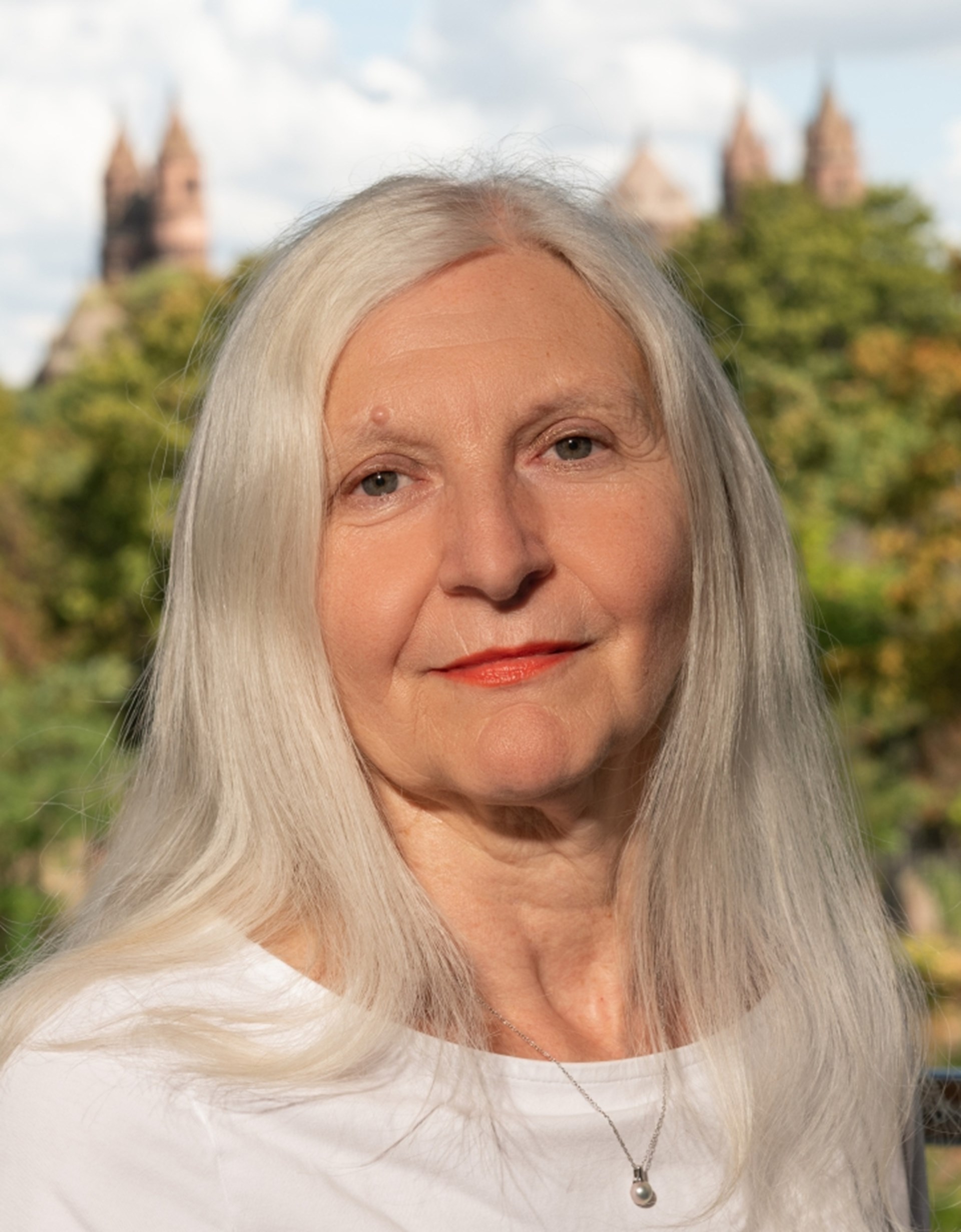 Porträtfoto einer Frau mit langen weiß-grauen Haaren in weißem Shirt; im Hintergrund unscharf grüne Bäume und die Silhouette des Wormser Doms