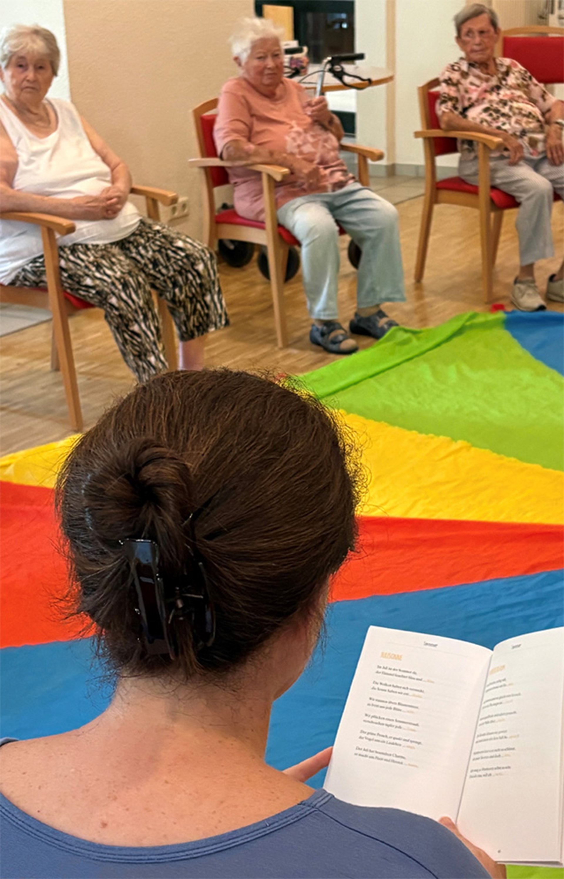 Rückansicht einer Person mit zusammengebundenen Haaren, die vor sich ein Buch in den Händen hält. Im Hintergrund sind drei ältere Personen, die auf Stühlen sitzen. Auf dem Boden vor den Stühlen liegt ein großes Tuch mit breiten farbigen Streifen in blau, rot, gelb und grün.