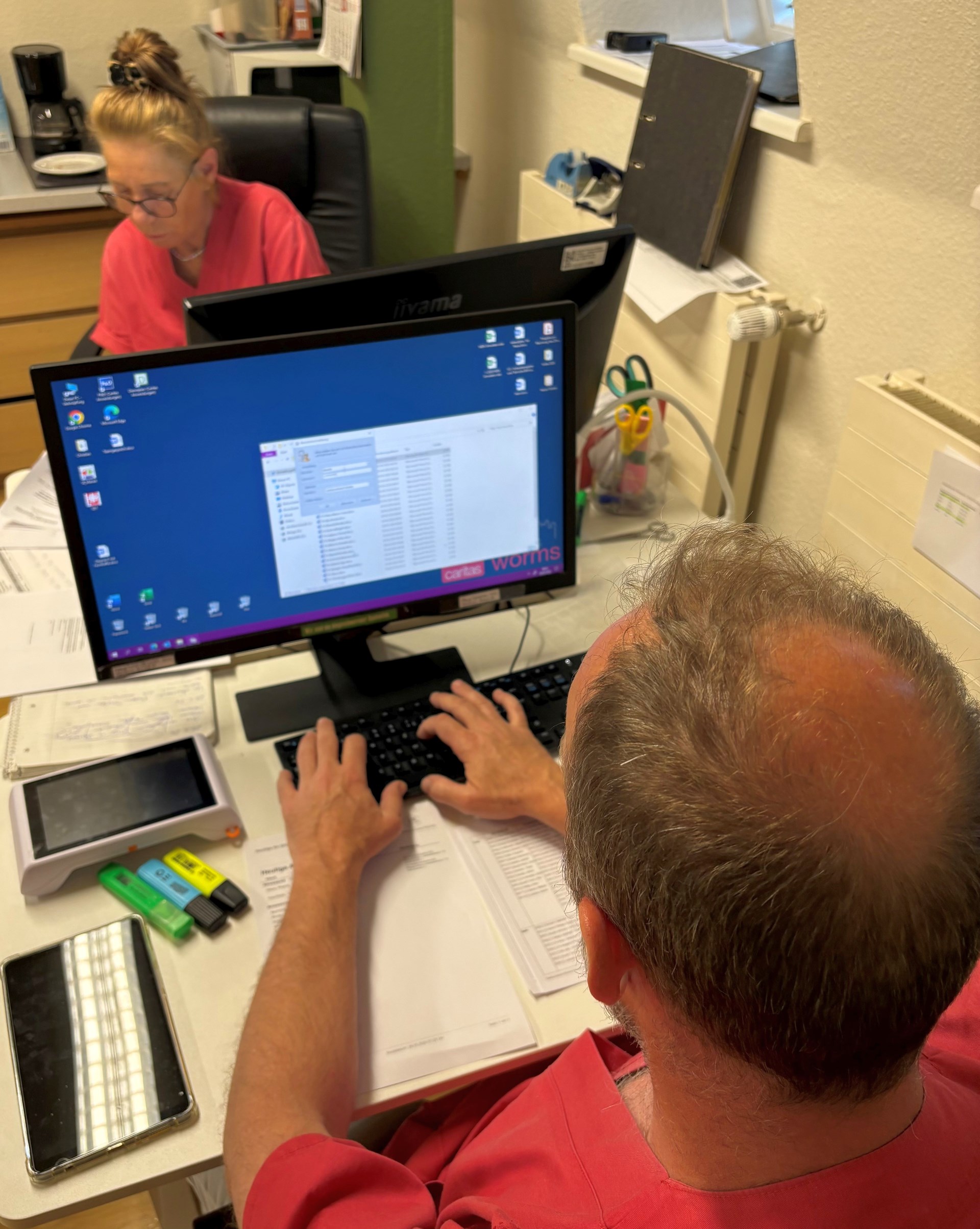 Zwei Mitarbeitende in einer kleinen Büro- bzw. Verwaltungsarbeitsumgebung. Im Vordergrund sitzt eine Person in roter Dienstkleidung am Schreibtisch und arbeitet an einer Tastatur vor einem Computerbildschirm mit geöffneten Dateien. Im Hintergrund sitzt eine weitere Person in pinker Dienstkleidung an einem Schreibtisch und arbeitet konzentriert. Auf dem Tisch liegen Büro- und Arbeitsmaterialien wie Marker, Unterlagen und ein Tablet.