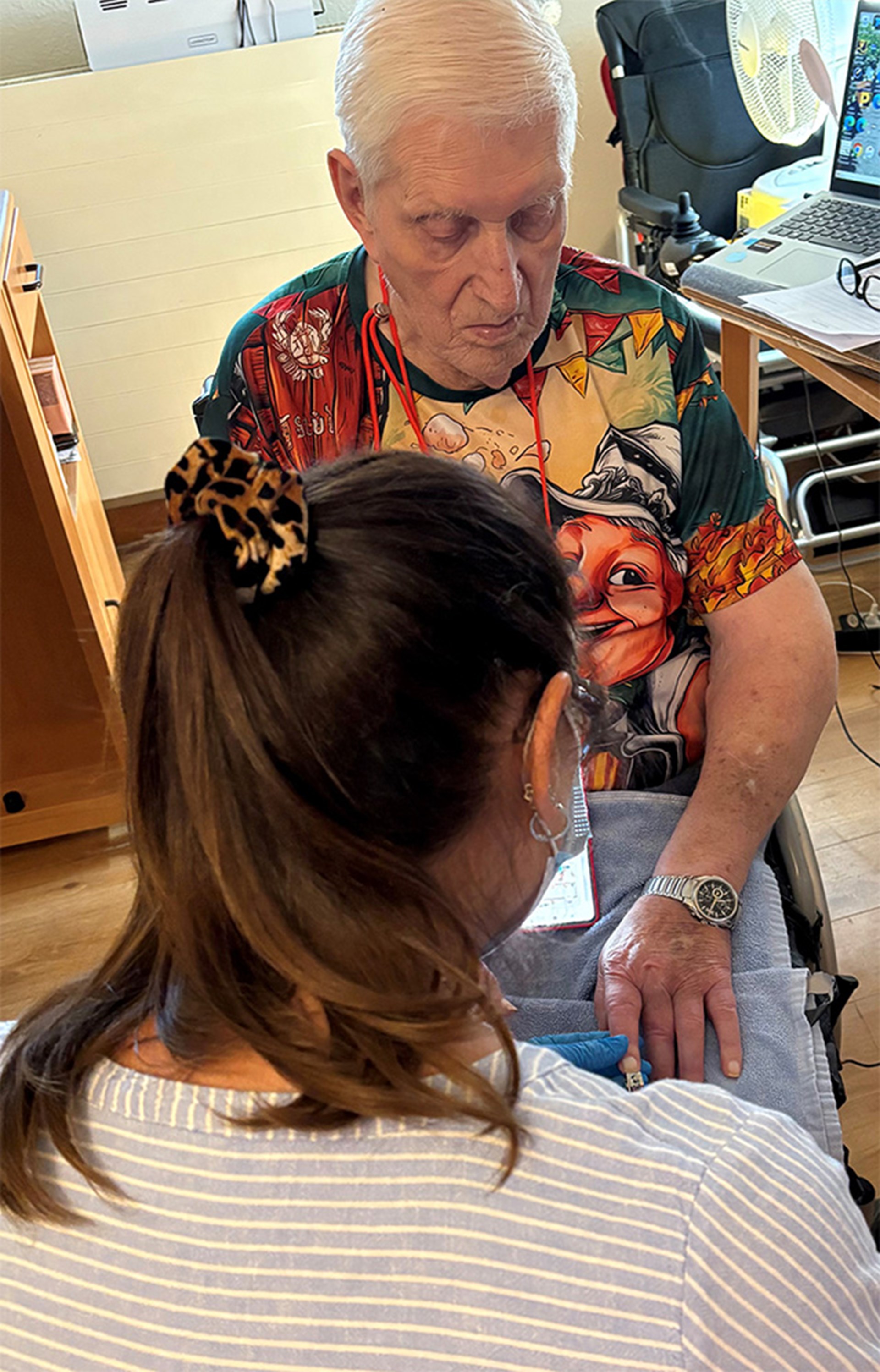 Rückansicht einer Person mit langem Zopf und geringeltem Oberteil. Dahinter sitzt eine Person, die auf die eigene Hand schaut, die von der Person im Vordergrund mit blauem Handschuh gehalten wird.