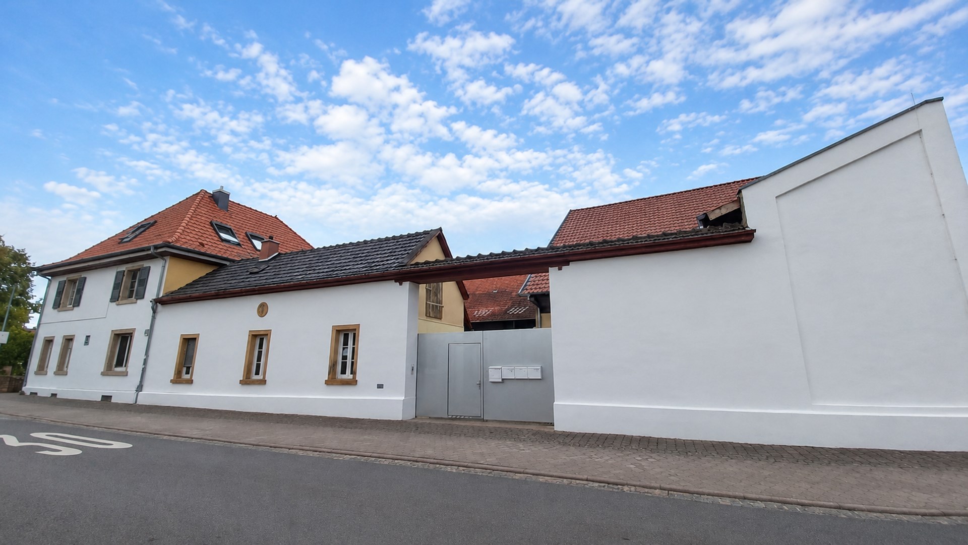 Straßenansicht eines hellen Gebäudekomplexes mit weißen Fassaden, roten Ziegeldächern und grauem Hoftor unter freundlichem, leicht bewölktem Himmel.