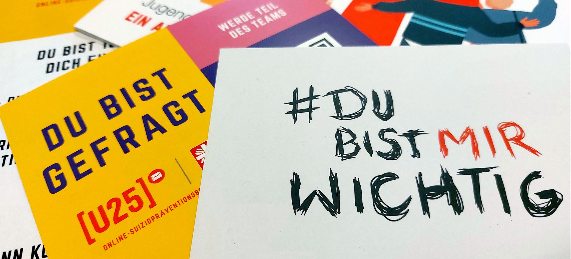 Mehrere bunte Informationskarten des Projekts U25 zur Online-Suizidprävention liegen auf einem Tisch. Auf einer gelben Karte steht „Du bist gefragt [U25]“. Im Vordergrund liegt eine weiße Karte mit handgeschriebenem Text: „#Du bist mir wichtig“, wobei „mir“ rot hervorgehoben ist.