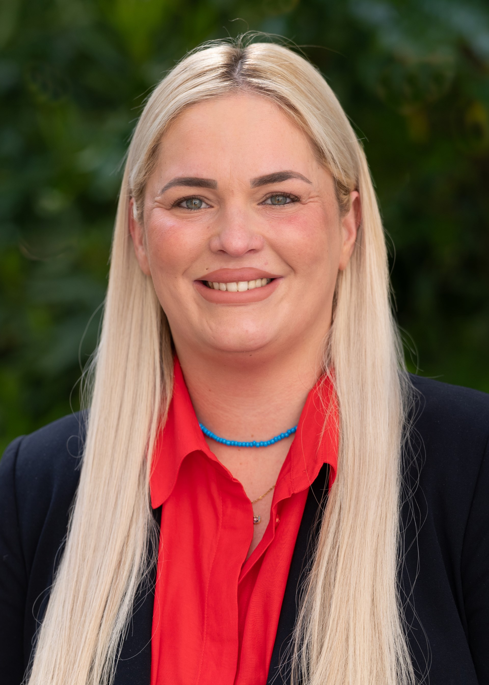 Porträt einer lächelnden Frau mit langen blonden Haaren. Sie trägt eine rote Bluse unter einem dunklen Blazer sowie eine türkisfarbene Halskette und blickt direkt in die Kamera. Im Hintergrund sind unscharfe grüne Pflanzen zu sehen. Die Aufnahme vermittelt einen freundlichen und professionellen Eindruck.