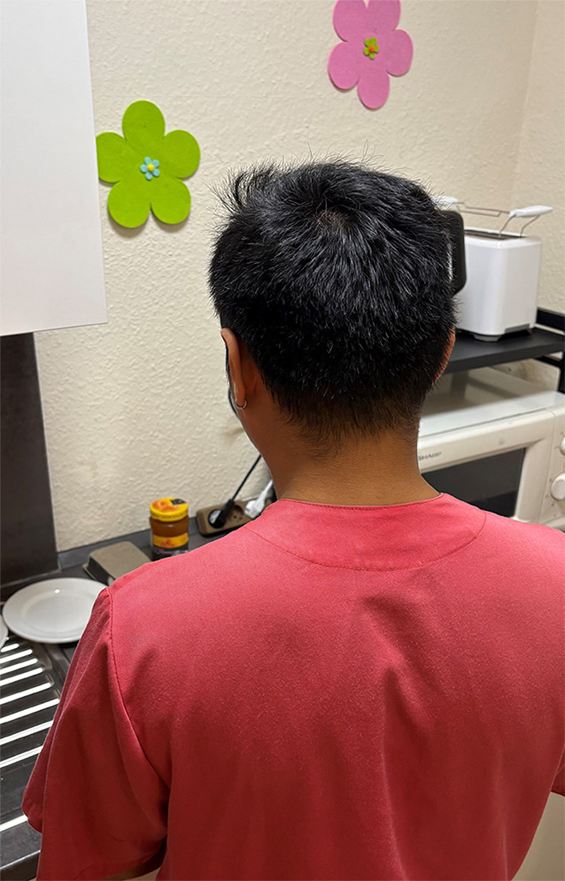 Rückansicht einer Pflegekraft in roter Arbeitskleidung in einer Küche. An der Wand hängen grüne und rosa Blütendekorationen. Auf der Arbeitsfläche stehen Küchengeräte und Geschirr. 