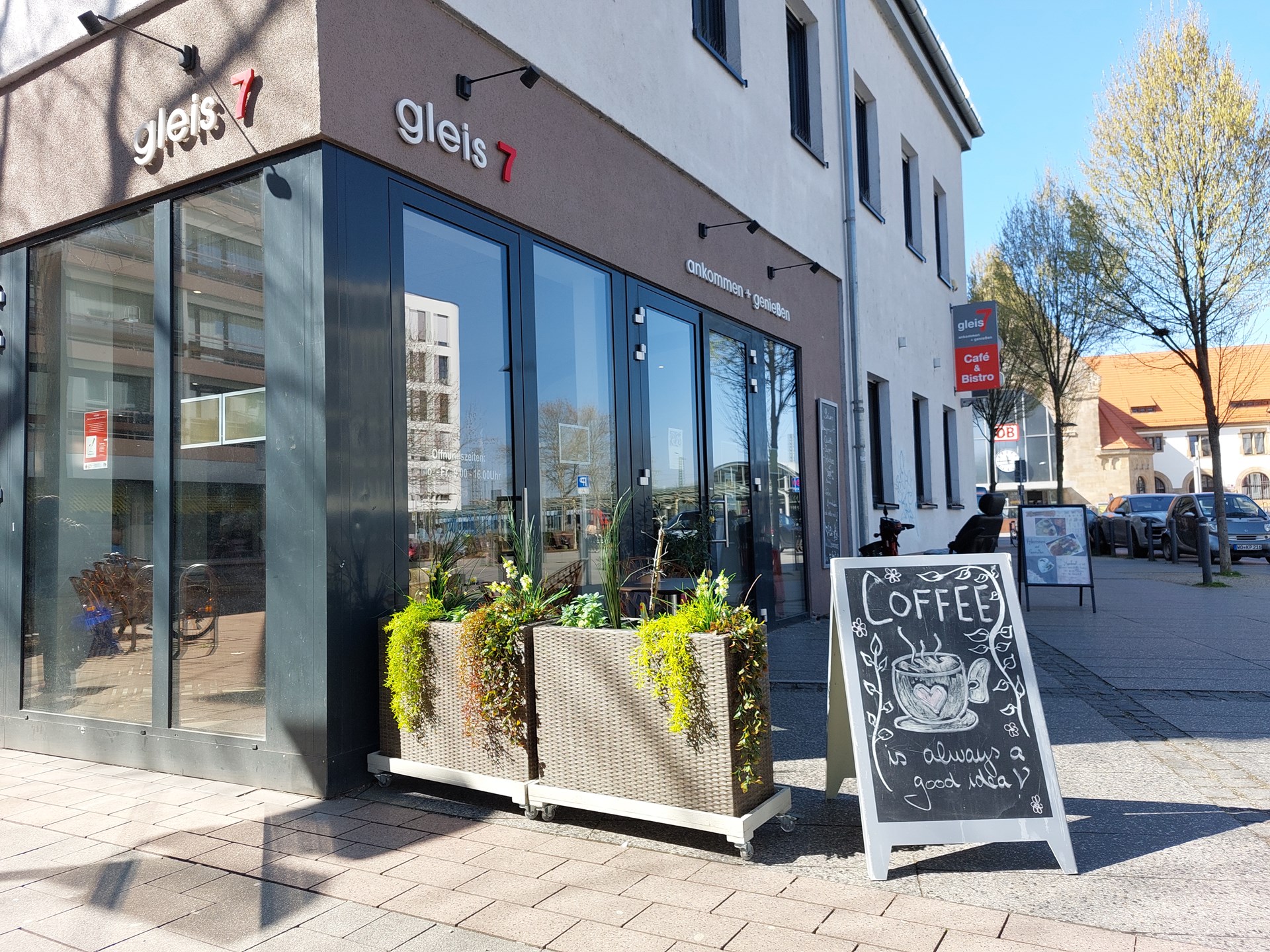 Außenaufnahme des Cafés „gleis 7“ mit großer Fensterfront und bepflanzten Kästen davor. Auf dem Gehweg steht eine Kreidetafel mit der Aufschrift „Coffee is always a good idea“ und einer Zeichnung einer Kaffeetasse. Entlang der Straße stehen weitere Schilder, Bäume und parkende Autos.