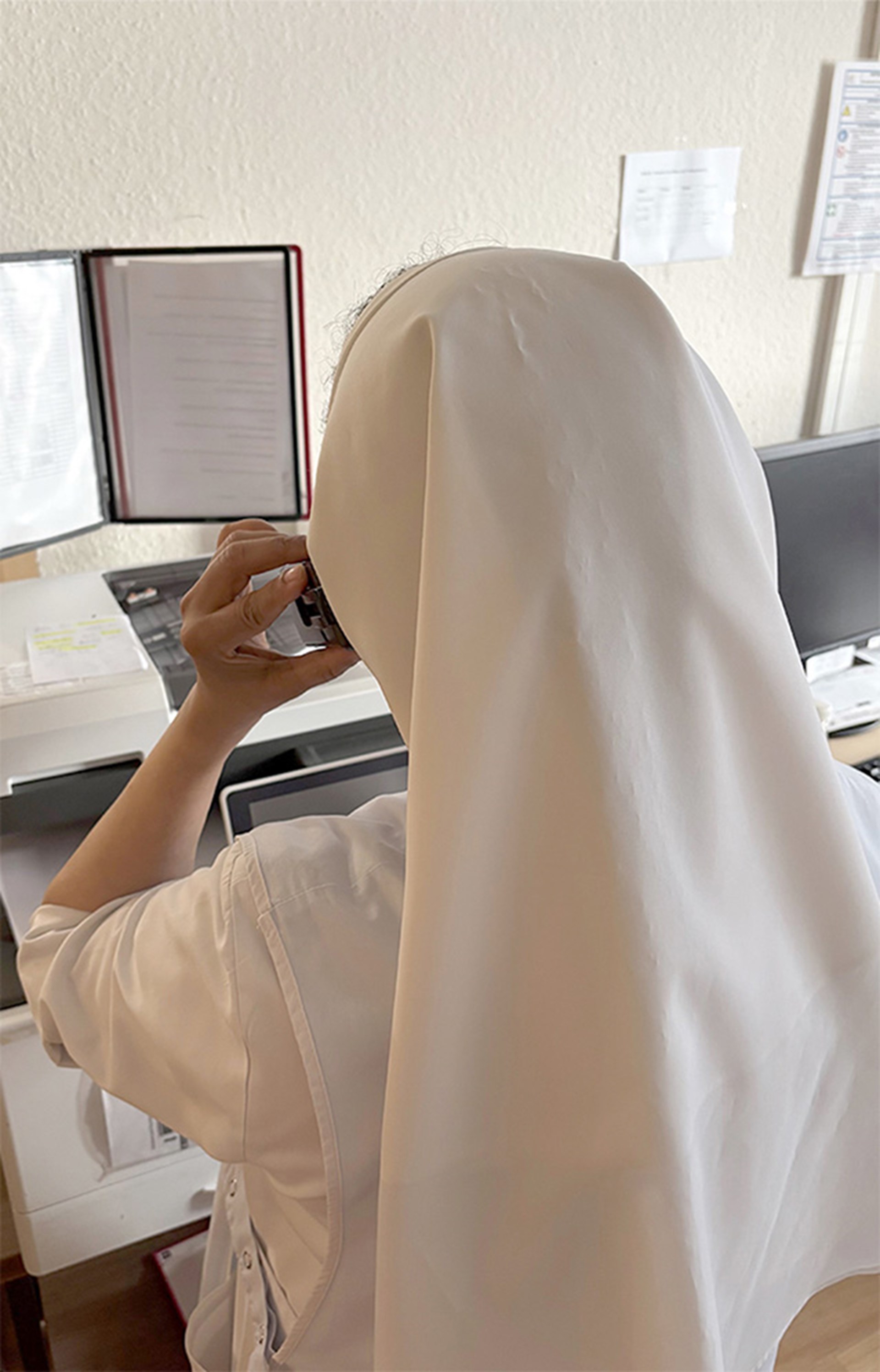 Rückansicht einer Pflegekraft in weißer Arbeitskleidung und Kopfbedeckung, die an einem Schreibtisch in einem Büro telefoniert. Auf dem Schreibtisch stehen ein Monitor und Arbeitsunterlagen. 