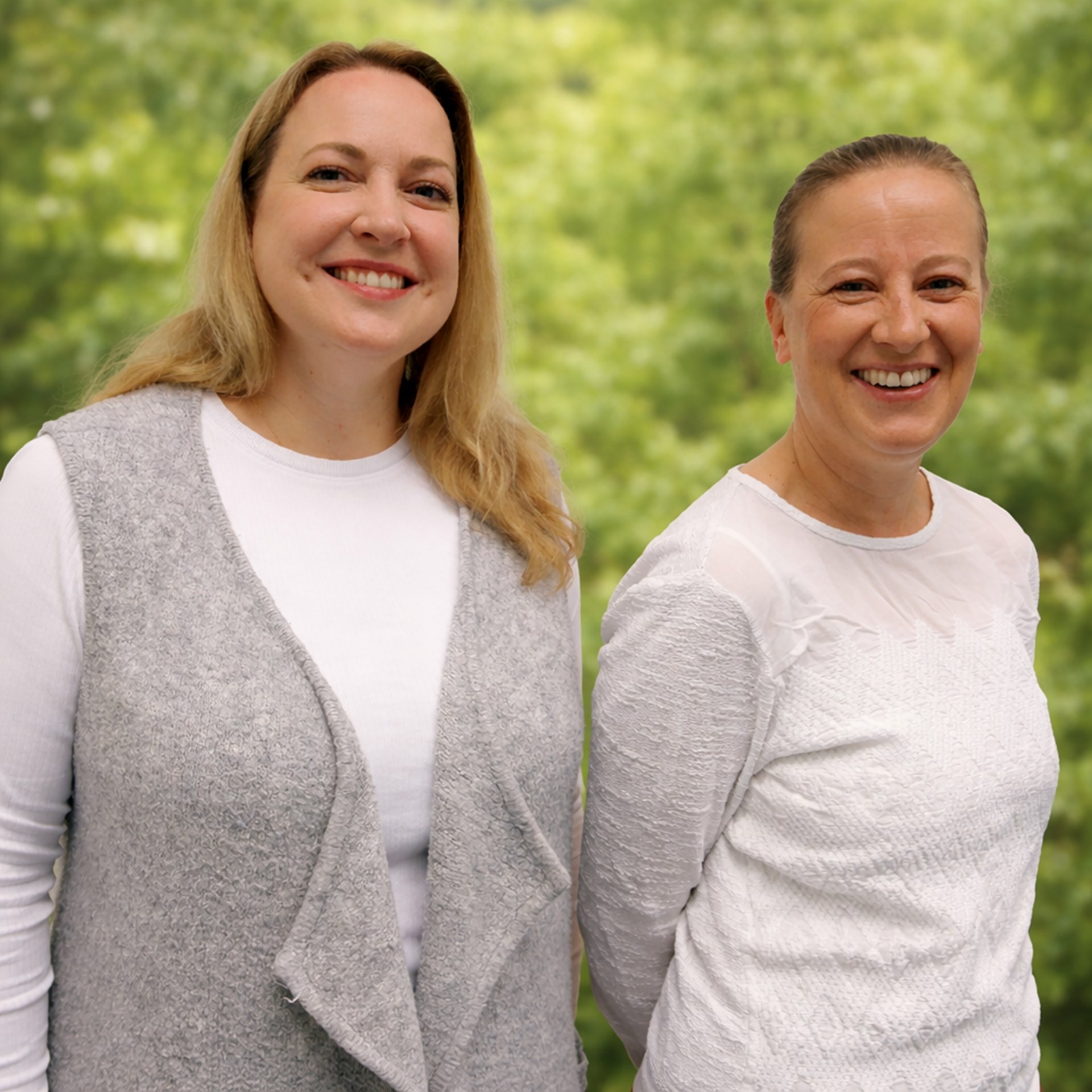 Zwei Frauen stehen nebeneinander, beide lächeln. Sie tragen helle, bequeme Kleidung. Im Hintergrund ist unscharf ein grünes Landschaftsbild zu sehen.
