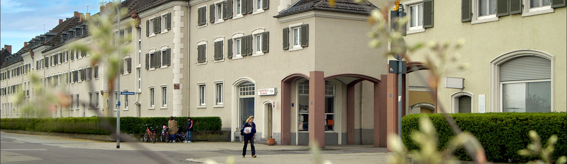 Außenansicht des Stadtteilbüros Nordend in Worms in einer langen, hellen Häuserzeile. Vor dem Gebäude sind mehrere Personen sowie Fahrräder zu sehen. Unscharfe Zweige und Blätter im Vordergrund rahmen die Aufnahme ein.