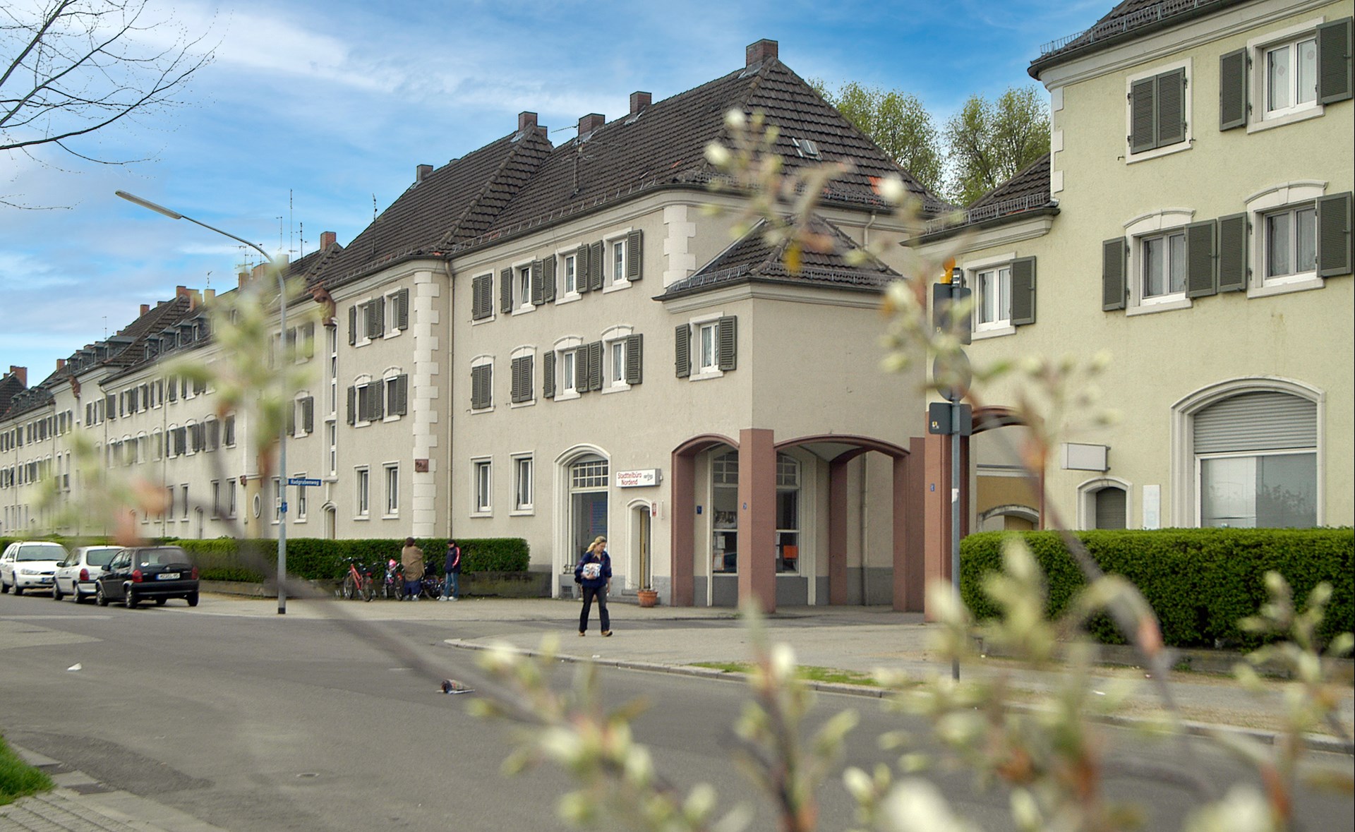 Außenansicht eines Caritas-Gebäudes in Worms mit mehreren verbundenen Wohn- und Geschäftshäusern entlang einer Straße. Vor dem Eingang stehen einige Menschen, im Vordergrund sind unscharfe Zweige zu sehen